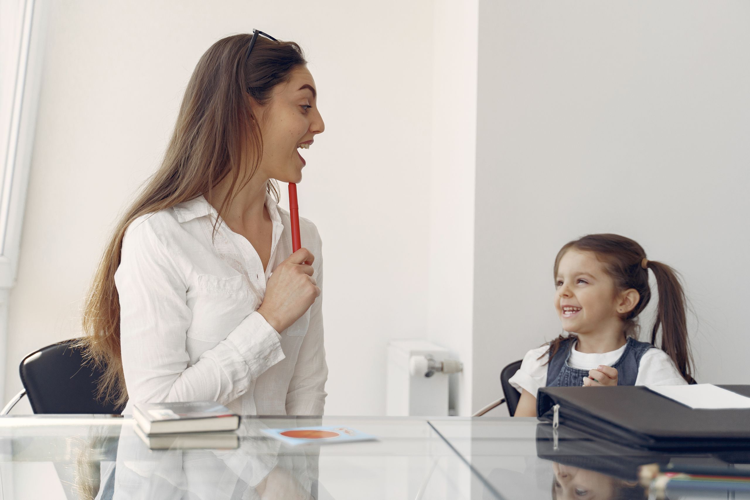 Positive female looking after little girl while sitting together at glass table in contemporary workplace and playng